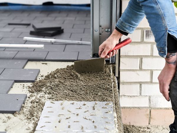 Handwerker bearbeitet mit einer Kelle frischen Mörtel am Rand einer teilweise verlegten dunklen Bodenplattenfläche.