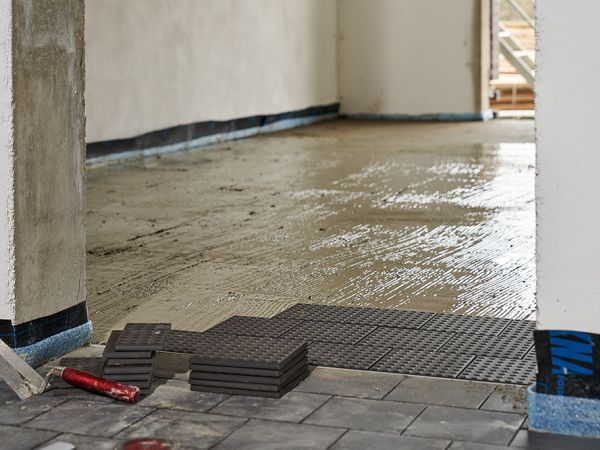 Baustelle mit vorbereitetem Mörtelbereich, gestapelten dunklen keramischen Bodenplatten und bereits verlegtem Bodenabschnitt.
